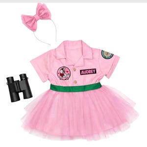 May include: A pink dress with a green waistband, a collar, and short sleeves. The dress features a patch of a dinosaur holding a heart, a patch with the name AUDREY, and a circular sleeve patch. Includes a pink bow headband and black binoculars.