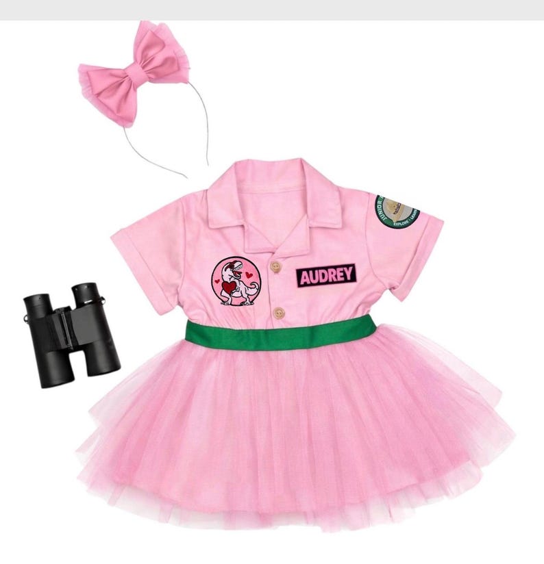 May include: A pink dress with a green waistband, a collar, and short sleeves. The dress features a patch of a dinosaur holding a heart, a patch with the name AUDREY, and a circular sleeve patch. Includes a pink bow headband and black binoculars.