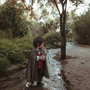 Puede incluir: Un ni&ntilde;o con una capa con capucha de color verde oliva oscuro sobre una camisa blanca de cuello, un chaleco rojo abotonado y pantalones de color marr&oacute;n oscuro. La capa se sujeta al cuello con un broche decorativo con una piedra verde.