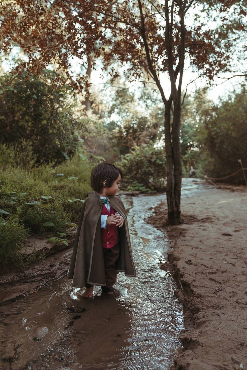 Puede incluir: Un ni&ntilde;o con una capa con capucha de color verde oliva oscuro sobre una camisa blanca de cuello, un chaleco rojo abotonado y pantalones de color marr&oacute;n oscuro. La capa se sujeta al cuello con un broche decorativo con una piedra verde.