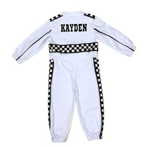 May include: A white long-sleeve jumpsuit featuring black and white checkered racing stripes along the sides and waist. The back displays the name KAYDEN in bold black block letters. The garment includes long sleeves with elastic cuffs and a collar.