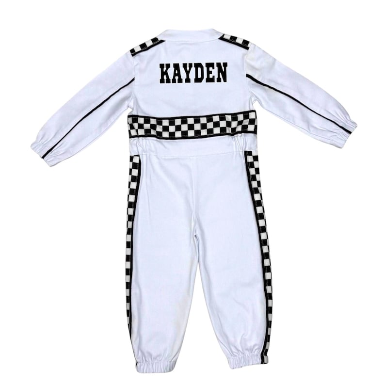 May include: A white long-sleeve jumpsuit featuring black and white checkered racing stripes along the sides and waist. The back displays the name KAYDEN in bold black block letters. The garment includes long sleeves with elastic cuffs and a collar.