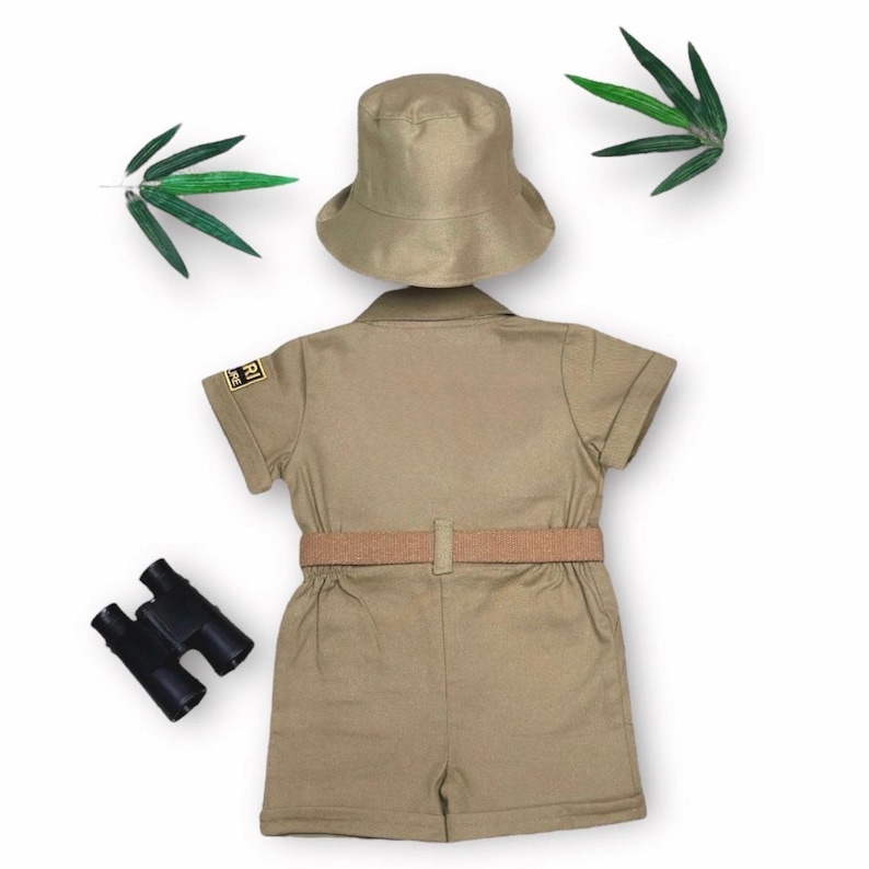 May include: A khaki-colored safari outfit for children, including a short-sleeved jumpsuit with a belt, a matching hat, and a pair of black binoculars. The outfit has a patch on the sleeve. The ensemble is designed for imaginative play and adventure.