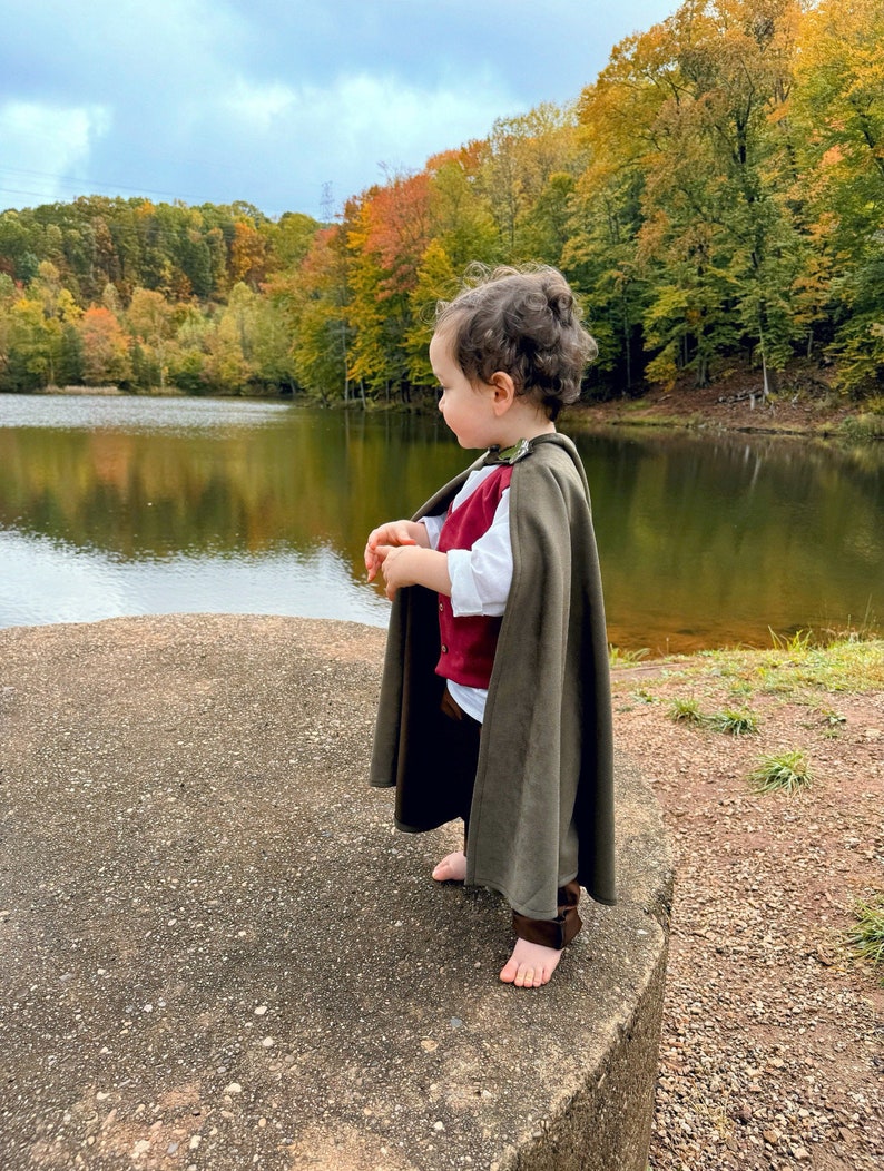 Puede incluir: Un conjunto para ni&ntilde;o peque&ntilde;o que consta de una capa con capucha de color verde oliva oscuro, un chaleco abotonado de color rojo intenso, una camisa blanca de manga larga y pantalones de color marr&oacute;n oscuro. La capa se sujeta al cuello.