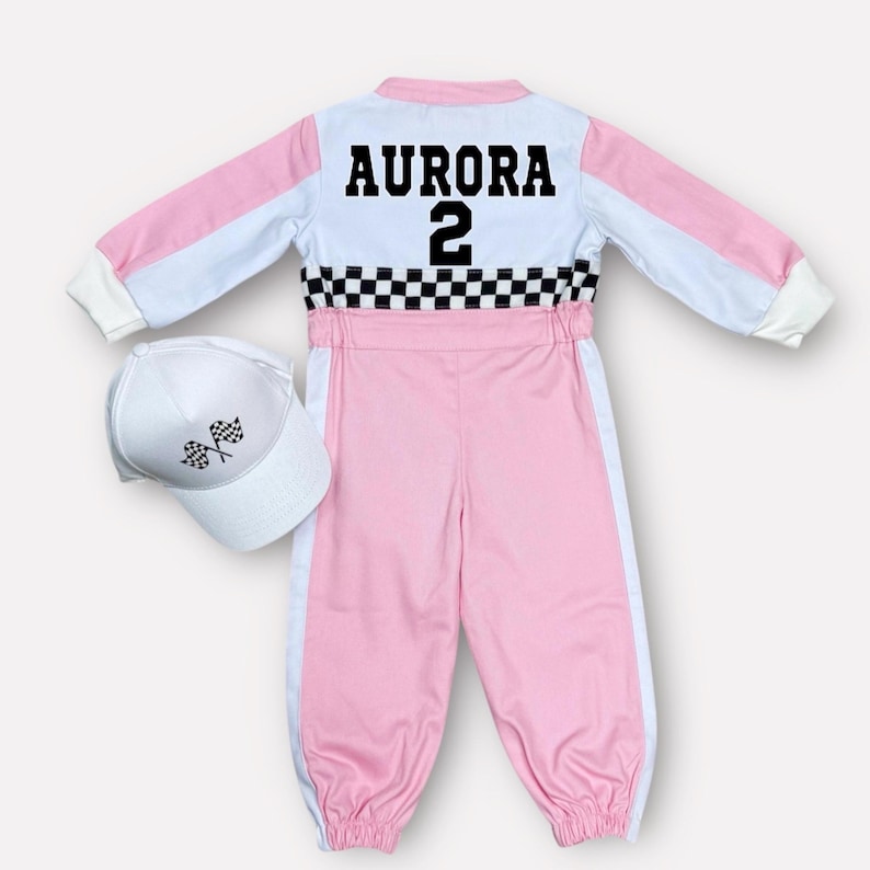 Op de afbeelding: Een roze en wit racepak met de naam "AURORA" en het nummer "2" op de rug. Een bijpassende witte baseballcap met geblokte vlaggen is inbegrepen. Het pak heeft een geblokt patroon in de taille.