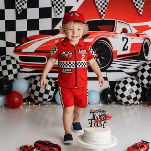 May include: A red racing-themed outfit consisting of a short-sleeved collared shirt with a zipper, matching shorts, and a baseball cap. The shirt features a checkered pattern, the name LIAM, the number 2, and a cartoon car graphic.