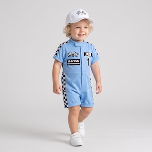 May include: A light blue short-sleeve racing romper with black and white checkered side panels, featuring the text JAKE, RACING, and a checkered flag graphic on the chest. The outfit includes a matching white baseball cap with a checkered flag logo.