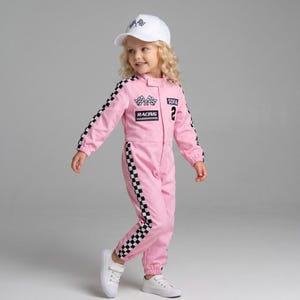May include: A pink racing jumpsuit featuring black and white chequered stripes down the sleeves and legs, with patches displaying the text SOFIA, 2, RACING, and chequered flags. The outfit is paired with a white baseball cap with a chequered flag logo.