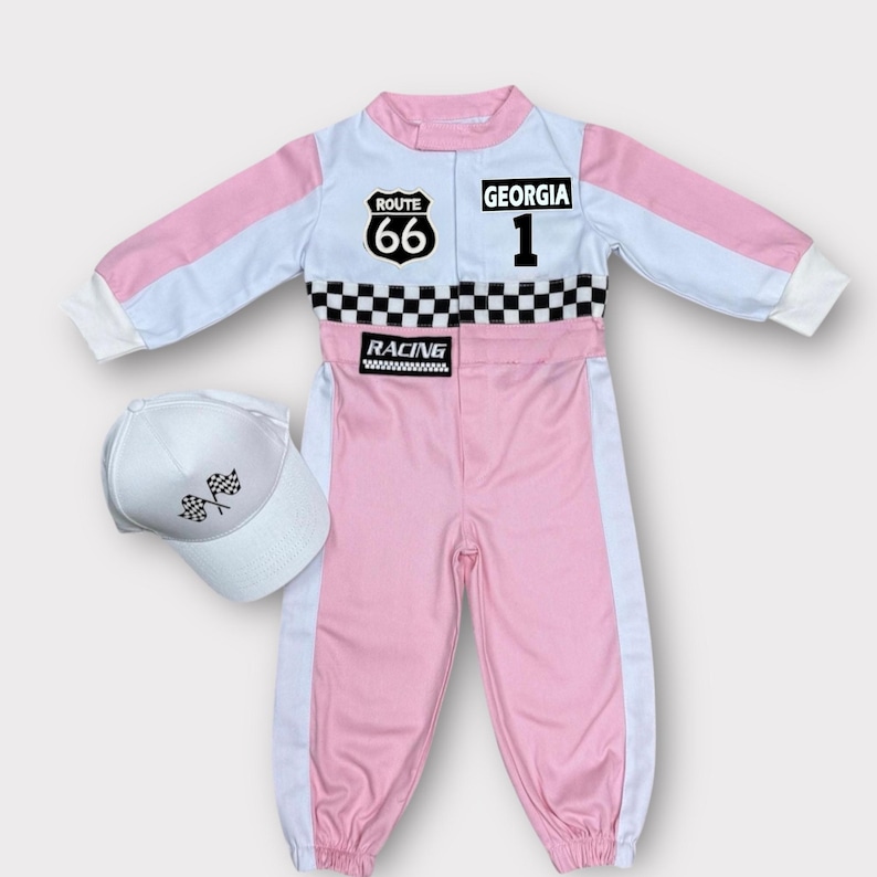 Op de afbeelding: Een roze, witte en zwarte race-overall met bijpassende witte pet. De overall heeft patches met "ROUTE 66" en "GEORGIA 1", een geblokte streep en de tekst "RACING". De pet heeft twee gekruiste geblokte vlaggen.