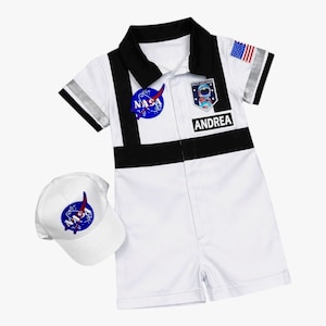 May include: White astronaut costume with black accents, a NASA patch, and the name "ANDREA". Includes a matching white baseball cap with a NASA logo. The costume has a zipper closure and short sleeves with silver stripes.