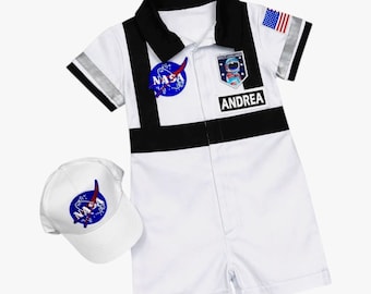 May include: A white short-sleeved romper with a black collar and black vertical stripes, featuring a blue NASA logo, a Space Explorer patch, a US flag patch, and a black name tag reading ANDREA. Includes a matching white baseball cap with a NASA logo.