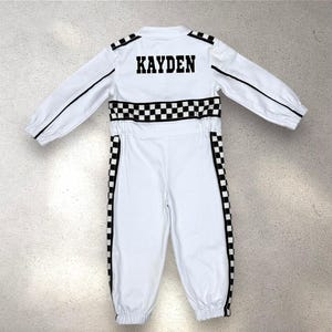 May include: White racing jumpsuit with black accents and the name "KAYDEN" on the back. The jumpsuit features a checkered pattern around the waist and down the sides, with black piping along the sleeves and legs. A stylish outfit for a young racer.