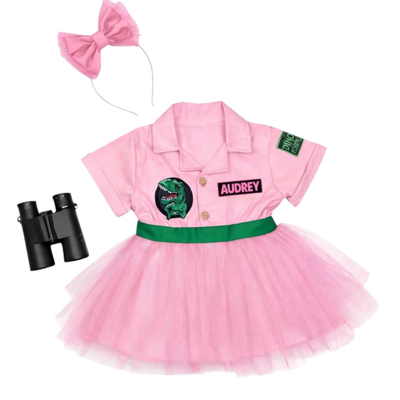 May include: A pink dinosaur explorer costume with a matching bow headband and black binoculars. The costume includes a collared shirt with a green dinosaur patch and the name "Audrey", a green belt, and a pink tutu skirt.