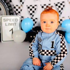 May include: A light blue racing-style jumpsuit featuring black and white checkered patterns on the sleeves and waist, with the number 1 in black on the chest. The garment includes a small checkered flag patch and a rectangular patch on the front.