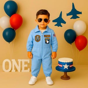 Könnte beinhalten: Ein Kind in einem hellblauen Air Force-Overall mit Aufnähern und Sonnenbrille steht vor Luftballons und einem Kuchen. Der Kuchen ist mit einem Stern und Streifen verziert. Das Wort "ONE" wird angezeigt.