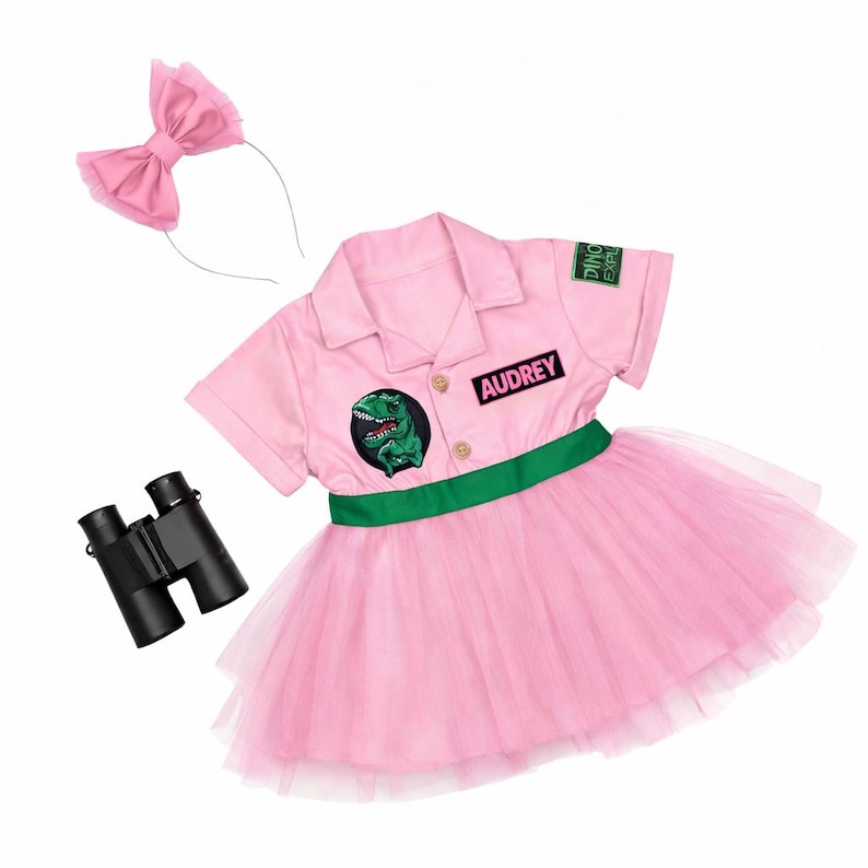 May include: Pink dinosaur explorer costume set with a collared shirt, tutu skirt, headband, and binoculars. The shirt has a green dinosaur patch and the name "Audrey". The costume includes a green belt and a pink bow.