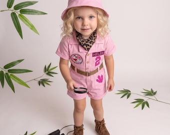 Puede incluir: Un mameluco rosa claro de manga corta con un sombrero de pescador a juego, pañuelo con estampado de leopardo, cinturón marrón, parche de dinosaurio, huella rosa y etiqueta con el nombre HELEN y el número 2, junto con botas marrones.