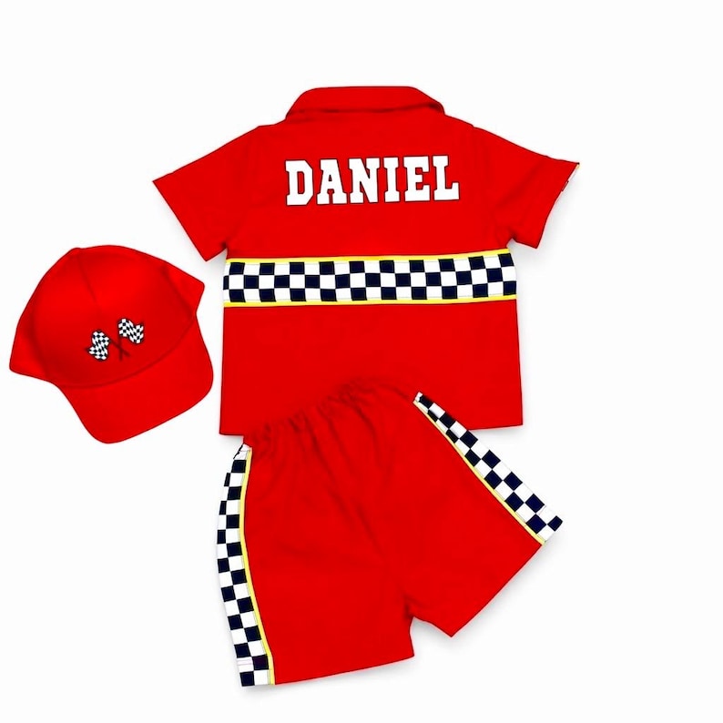 May include: A red short-sleeved button-down shirt with the name DANIEL printed in white, paired with matching red shorts and a red baseball cap. The shirt and shorts feature a black and white checkered racing stripe. The cap has two checkered flags.