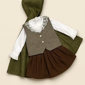 May include: A children's outfit comprising a white blouse with lace trim, a grey waistcoat with two buttons, and a brown pleated skirt. A green hooded cape is draped behind the outfit.