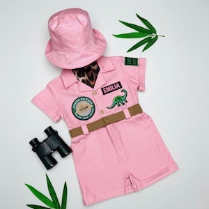 May include: A pink bucket hat and a matching pink short-sleeve romper featuring a brown belt, a Junior Ranger patch, a green dinosaur patch, and a black name tag reading EMILIA. Black binoculars are included with the outfit set.