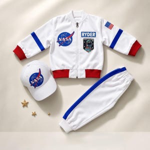 Può includere: Un completo bianco a tema NASA composto da un giubbotto bomber con zip, dettagli rossi e blu, pantaloni della tuta bianchi abbinati con strisce laterali blu e un cappellino da baseball bianco con il logo della NASA e la scritta RYDER.