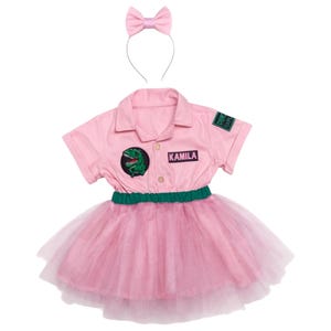 May include: A pink dinosaur-themed costume set. The set includes a short-sleeved collared shirt with a dinosaur patch and the name "KAMILA", a tutu skirt, and a matching bow headband. The shirt has a green and black patch that says "Dino Exp".