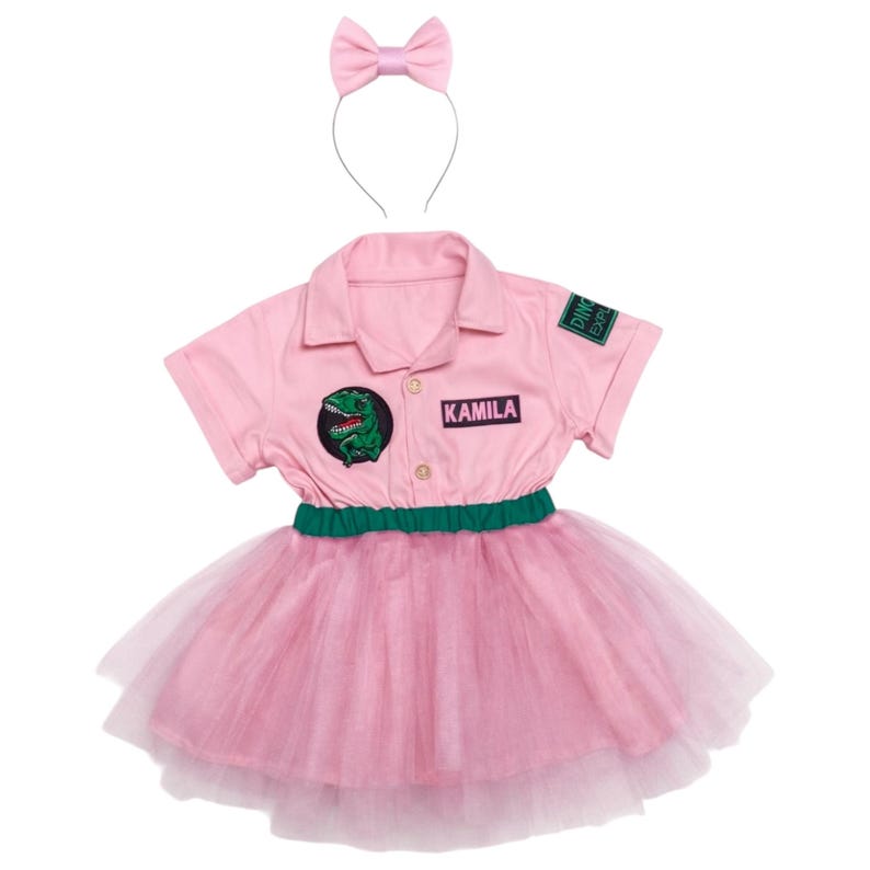 May include: A pink dinosaur-themed costume set. The set includes a short-sleeved collared shirt with a dinosaur patch and the name "KAMILA", a tutu skirt, and a matching bow headband. The shirt has a green and black patch that says "Dino Exp".