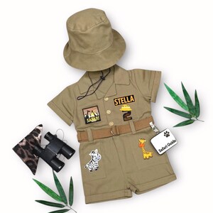 May include: Khaki safari-themed outfit set with a matching hat, shorts, and shirt. The shirt has the name "STELLA" and a "Safari" patch. Includes a belt, binoculars, and a "Safari Guide" tag. Features zebra and giraffe patches.