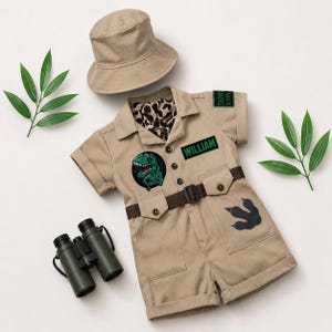 May include: A tan short-sleeved romper with a button-down front, a brown belt, and a bucket hat. The romper features a green dinosaur patch, a black patch with the name WILLIAM, a DINO EXPLORER patch, and a black dinosaur footprint graphic.