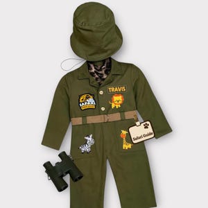 Puede incluir: Conjunto de disfraz de safari verde oliva con sombrero, cinturón y prismáticos a juego. El mono presenta parches de animales, una etiqueta con el nombre "Travis" y una etiqueta de "Guía de safari". El disfraz incluye una bufanda con estampado de leopardo.