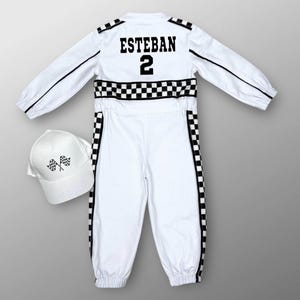 May include: White racing suit with black checkered accents and the name "ESTEBAN" and the number "2" on the back. Includes a matching white baseball cap with checkered flags. The suit has black stripes down the sleeves and legs.