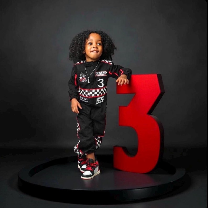 Puede incluir: Un ni&ntilde;o con un mono de carreras negro con detalles rojos y blancos, el n&uacute;mero 3 y la palabra "LEGEND" en el pecho. El ni&ntilde;o est&aacute; de pie junto a un gran n&uacute;mero 3 rojo. Lleva zapatillas deportivas negras, rojas y blancas.