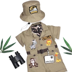 May include: A tan safari-themed outfit consisting of a short-sleeved romper and a matching bucket hat. The romper features a leopard print collar, a brown belt, and patches of a zebra, lion, monkey, giraffe, and a name tag reading STELLA.