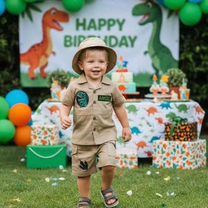 Pode incluir: Um conjunto de camisa e calções estilo safari em tom bege com um chapéu de pescador a combinar. A camisa apresenta um emblema de dinossauro verde no peito e um emblema retangular com o nome ANGEL. Os calções incluem um gráfico de pegada preta.