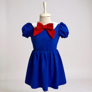 May include: A blue dress with a white collar and a large red bow. The dress has short, puffed sleeves and a gathered skirt. The dress is displayed on a mannequin.