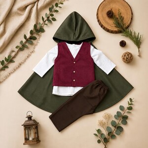 Puede incluir: Un conjunto que consta de una capa con capucha de color verde oscuro, una camisa blanca de manga larga, un chaleco burdeos con botones y pantalones de color marr&oacute;n oscuro. El conjunto est&aacute; dispuesto en plano, mostrando los componentes de la prenda.