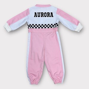 Op de afbeelding: Een roze en wit racepak met de naam "AURORA" in zwarte blokletters op de rug. Het pak heeft een geblokt patroon in de taille en witte strepen aan de zijkanten.