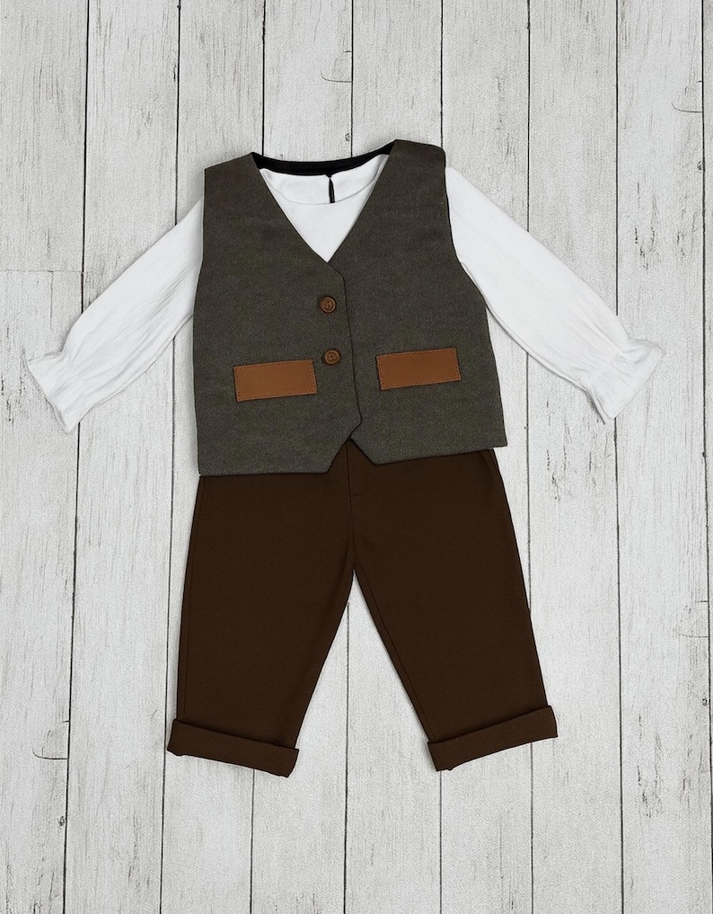 May include: A long-sleeved white shirt with ruffled cuffs paired with a dark gray vest featuring two brown rectangular pockets and two brown buttons. The outfit includes solid dark brown trousers with cuffed hems, creating a complete set.