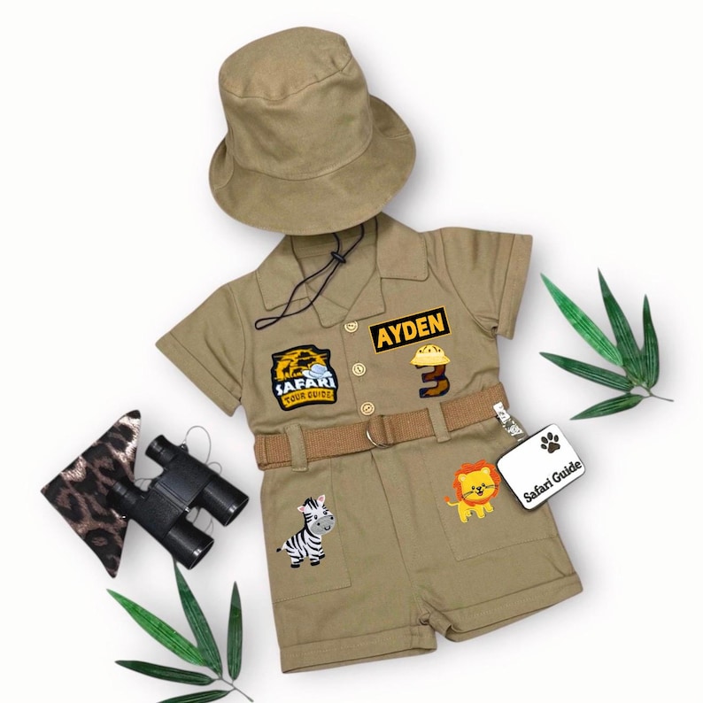May include: A khaki safari-themed outfit for a child, including a short-sleeved shirt and shorts set, a matching hat, and a belt. The shirt has patches that say "Safari Tour Guide" and "Ayden". The shorts have patches of a zebra and a lion. Includes binoculars and a leopard print accessory.