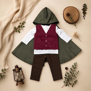 Puede incluir: Un conjunto de disfraz que consta de una capa con capucha de color verde oliva oscuro, una camisa blanca de manga larga, un chaleco de color burdeos intenso con tres botones blancos y pantalones de color marr&oacute;n oscuro, dispuestos en plano.