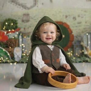 May include: A toddler wearing a dark green hooded cloak over a white long-sleeved shirt, a brown vest with two buttons, and brown pants. The toddler is holding a circular wooden ring with engraved text around the outer edge of the band.