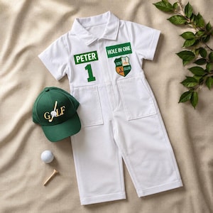 May include: A white short-sleeved romper featuring the text "PETER" and "HOLE IN ONE" with a green crest logo, paired with a dark green baseball cap embroidered with "GOLF" and a golf ball graphic, a white golf ball, and a wooden golf tee.