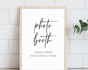 May include: A white sign with a black wooden frame, with the text "photo booth" in cursive script and the text "Grab a prop and strike a pose" in all caps.