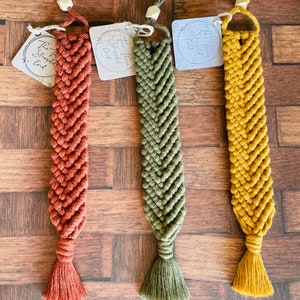 May include: Three macrame wall hangings with tassels in rust, olive green, and mustard yellow. Each hanging has a tag that reads "The Stranded Cord".