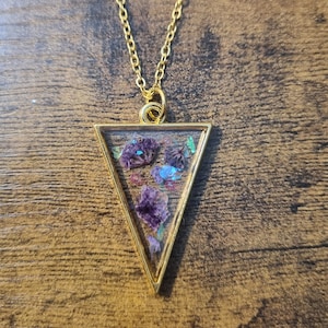 May include: A gold chain necklace with a triangular pendant featuring pressed purple flowers and iridescent glitter inside a clear resin.