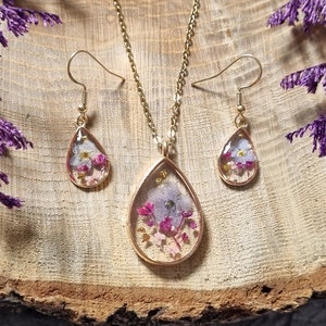 Peut inclure: Un collier et des boucles d'oreilles en or avec des pendentifs en forme de goutte d'eau remplis de fleurs pressées. Les fleurs sont un mélange de blanc, de rose et de violet.