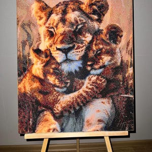 Peut inclure: Une peinture diamant représentant une lionne et son petit, dans des tons de marron, d'orange et de beige. L'œuvre est exposée sur un chevalet en bois, mettant en valeur la mosaïque détaillée de l'image. La lionne et son petit s'embrassent.