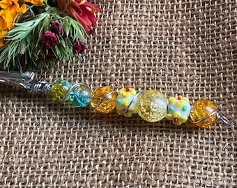 May include: A silver wire wrapped with colorful glass beads. The beads are yellow, white, blue, and red. The beads are arranged in a line and are separated by silver wire.