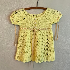 May include: A yellow crocheted dress with a scalloped hem and short sleeves. The dress has a ribbon tie at the waist and a button closure at the neckline.