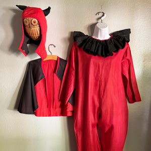 May include: A red clown costume with a black ruffled collar and matching pants. A red and black cape and a devil-horned hood with a carved face are also included. The costume is hanging on a wall.